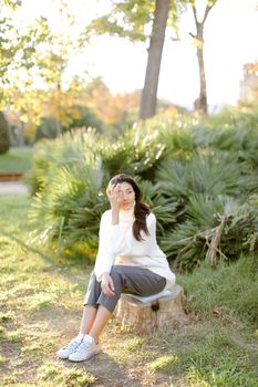 Chinese woman sitting on stump in park and wearing white sweater. Concept of asian female person.