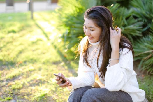 Japanese female student sitting in park and typing by smartphone. Concept of modern technology and social networks.