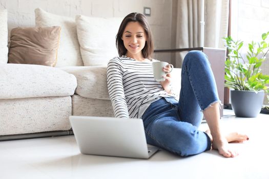 Pretty woman working with laptop while sitting on floor at home and drinking coffee
