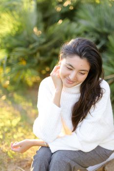 Japanese woman in white sweater sitting on grass in park. Concept of asian female person.