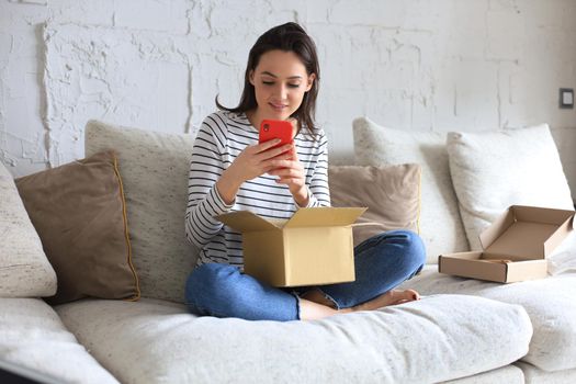 Beautiful young woman is holding cardboard box and unpacking smartphone sitting on sofa at home