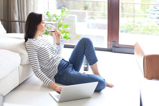 Smiling young woman sitting on floor with laptop computer and chating with friends, drinking wine