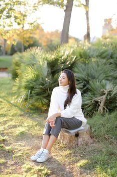 Japanese woman sitting on stump in park and wearing white sweater. Concept of asian female person.