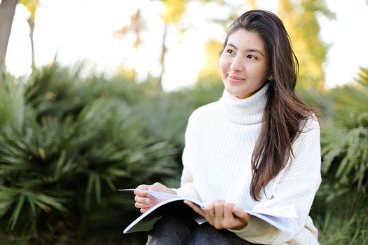 Chinese smiling female student sitting on stump in park and reading papers. Concept of preparing before exam and asian woman.