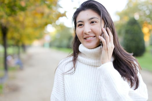 Asian pretty woman wearing white sweater speaking by smartphone walking in autumn park. Concept of chinese girl and modern technology.