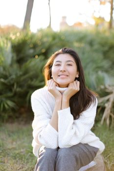 Chinese smiling woman sitting on stump in park and wearing white sweater. Concept of asian female person.