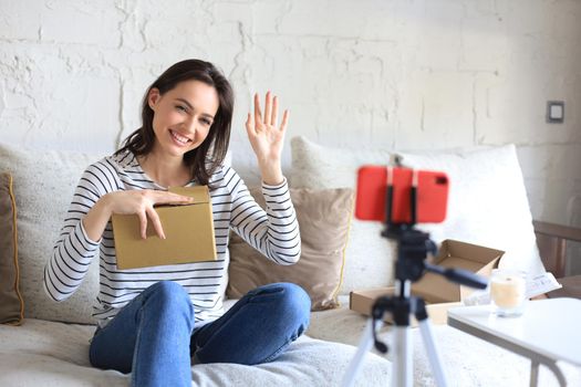 Young beautiful female blogger recording video while unpacking parcel at home