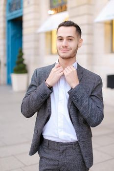 Young cacuasian male person wearing suit and wite shirt standing near building outdoors. Concept of fashion and businessman, urban life.