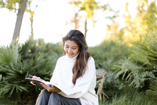 Chinese happy female student sitting on stump in park and reading papers. Concept of preparing before exam and asian woman.