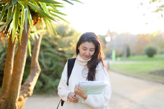 hinese girl with documents walking in park and wearing white sweater with backpack. Concept of asian student.