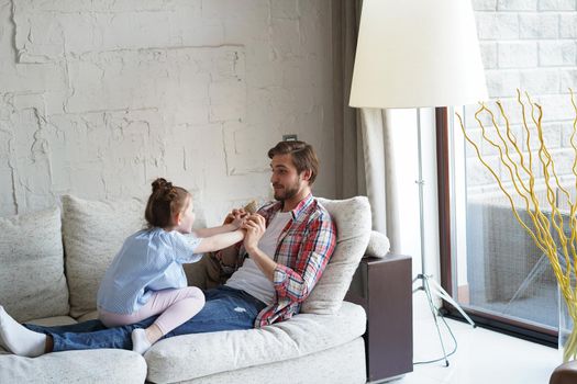 Cute little kid daughter laughing tickling playing with dad on sofa, happy father relaxing having fun with funny small child girl bonding enjoying leisure together in living room