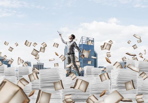 Man in casual wear keeping hand with book up while standing among flying books with cityscape on background. Mixed media.
