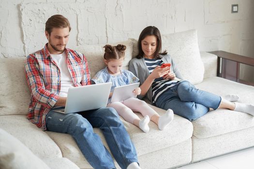 Father, mother and daughter using electronic devices sitting on sofa at living room