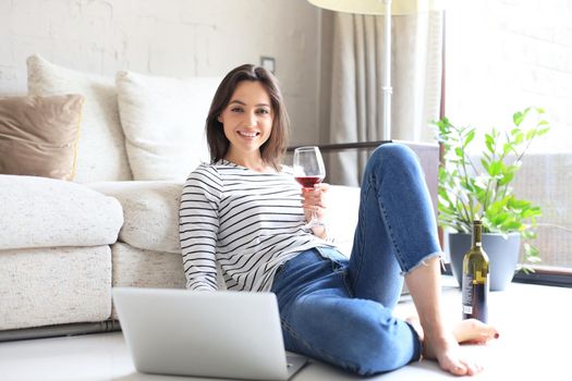 Smiling young woman sitting on floor with laptop computer and chating with friends, drinking wine