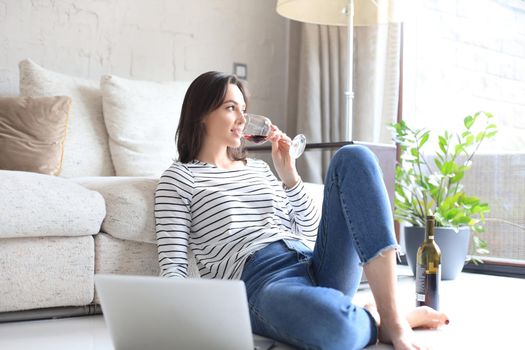 Smiling young woman sitting on floor with laptop computer and chating with friends, drinking wine