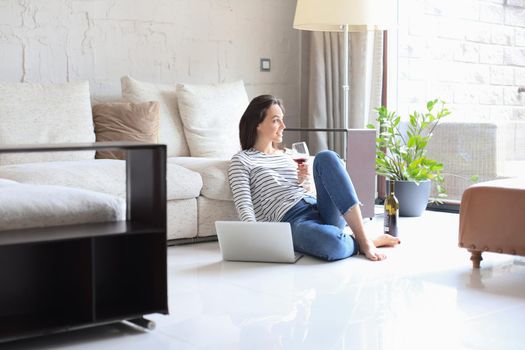 Smiling young woman sitting on floor with laptop computer and chating with friends, drinking wine