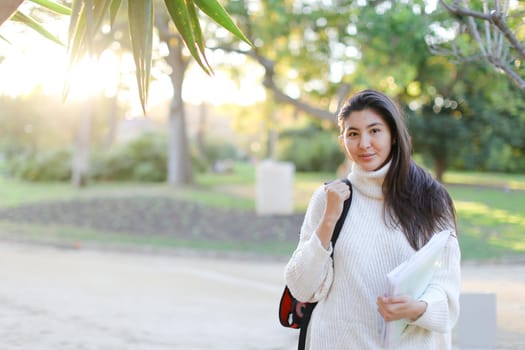 Korean girl with documents walking in park and wearing white sweater with backpack. Concept of asian student.