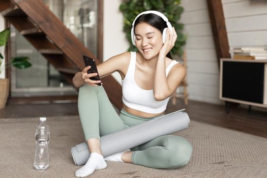 Happy sporty girl sitting on floor at home and taking selfie during workout, wearing headphones and holding smartphone. Concept of sport and lifestyle