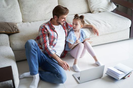 Smiling father and daughter sitting on floor in living room with laptop, teaching lessons