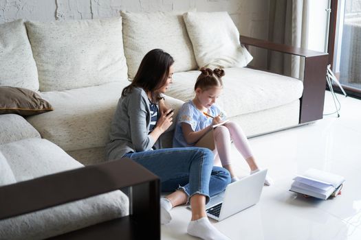 Smiling mother and daughter sitting on floor in living room with laptop, teaching lessons