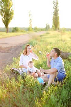 Young father and blonde nice mother sitting on grass with little baby and blowing bubbles. Concept of picnic with child and resting on nature.