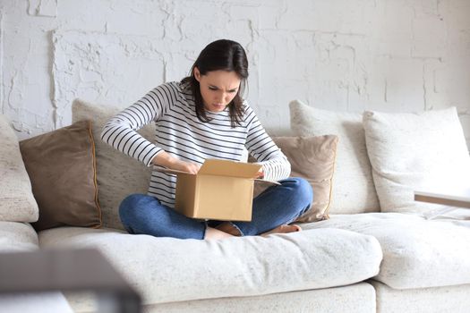 Beautiful young woman is holding cardboard box and unpacking it sitting on sofa at home