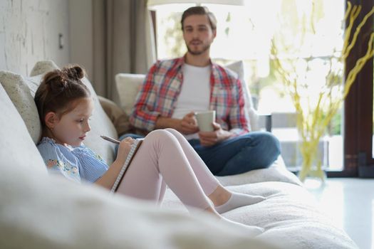 Happy father smilling daughter sitting on sofa enjoying creative activity, drawing pen pictures in albums, father and daughter spend free time together