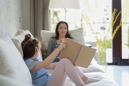 Happy mother smilling daughter sitting on sofa enjoying creative activity, drawing pen pictures in albums, mother and daughter spend free time together