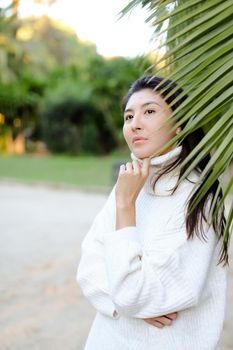 Chinese pretty woman in white sweater standing near palm leaf. Concept of asian beauty and female person on tropical resort.