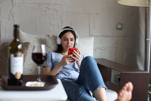 Pretty girl using her smartphone on couch at home in the living room. Listening music, drinking red wine, relaxation after a hard week at work