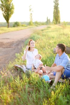 Young father and blonde mother sitting on grass with little baby and blowing bubbles. Concept of picnic with child and resting on nature.