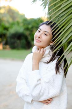 Chinese smiling woman in white sweater standing near palm leaf. Concept of asian beauty and female person on tropical resort.