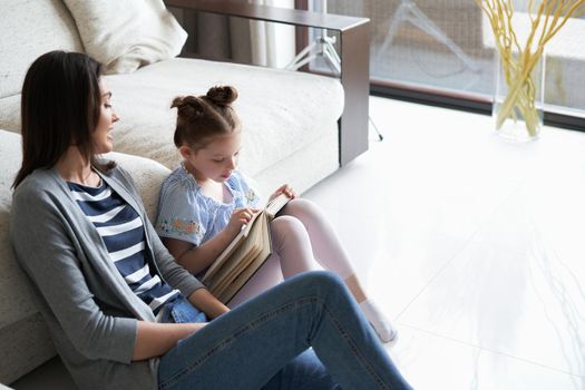 Smiling young mother and little daughter sitting on floor near sofa at home and reading books