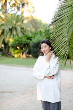 Korean cute woman in white sweater standing near palm leaf. Concept of asian beauty and female person on tropical resort.