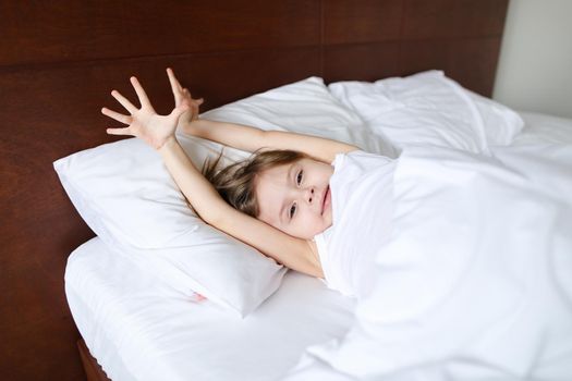 Little pretty girl with raised hands sleeping on bed with white linens in morning. Concept of childhood and resting.