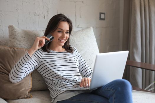 Happy natural brunette using credit card and laptop in the living room