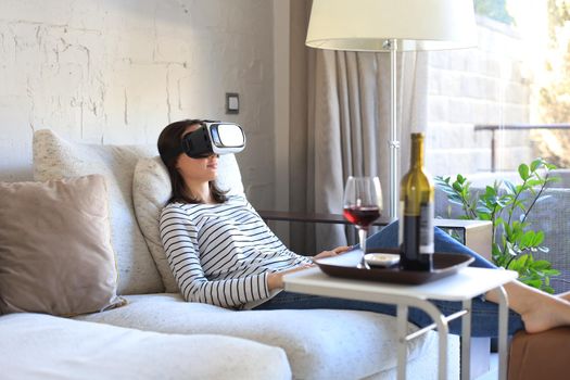 Young casual girl wearing VR headset while lounging on couch at home and looking around