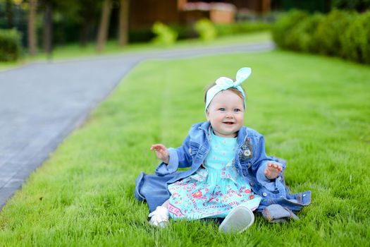 Little cute female baby sitting on green grass and wearing jeans jacket. Concept of childhood and resting in garden, nature background.