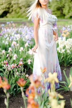 Young caucasian woman wearing white dress standing near irises on garden. Concept of human beauty and flora, spring inspiration.