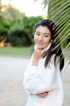 Chinese nice woman in white sweater standing near palm leaf. Concept of asian beauty and female person on tropical resort.