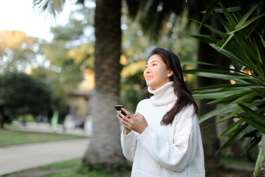 Chinese nice girl using smartphone and walking in tropical park. Concept of modern technology and nature.