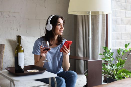 Pretty girl using her smartphone on couch at home in the living room. Listening music, drinking red wine, relaxation after a hard week at work