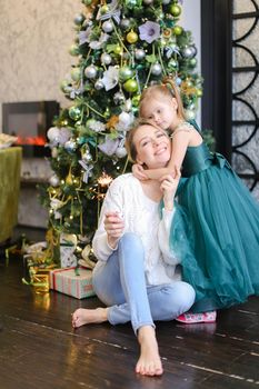 Young woman with bengal light wearing jeans and sitting with daughter near Christmas tree. Concept of celebrating winter holidays and positive emotions.