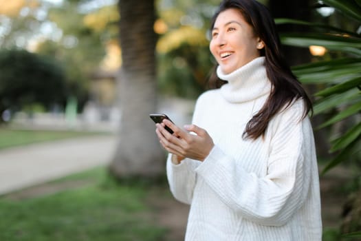 Focus on smartphone in chinese woman hands, girl standing in tropical park. Concept of modern technology and nature.