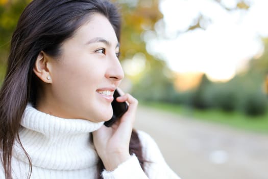 Close up asian smling woman wearing white sweater speaking by by smartphone walking in autumn park. Concept of chinese girl and modern technology.