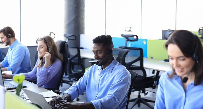 Smiling african male call-center operator with headphones sitting at modern office with collegues on the backgroung, consulting online