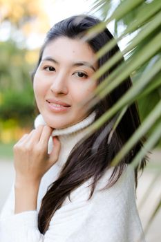 Portrait of chinese nice woman wearing white sweater standing near palm leaf. Concept of asian beauty and female person.