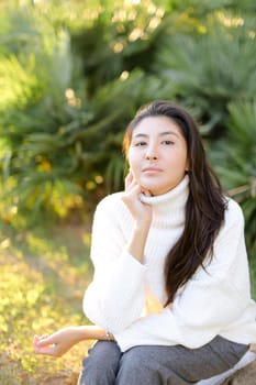 Korean woman in white sweater sitting on grass in park. Concept of asian female person.