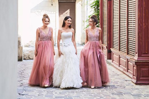 Beautiful bride and bridesmaids in gorgeous elegant stylish red pink violet floor length v neck chiffon gown dress decorated with sequins sparkles and rhinestones holding flowers bouquets. Wedding day in old beautiful European city.