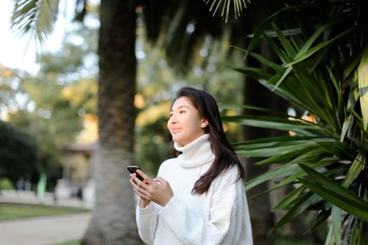 Chinese girl using smartphone and walking in tropical park. Concept of modern technology and nature.